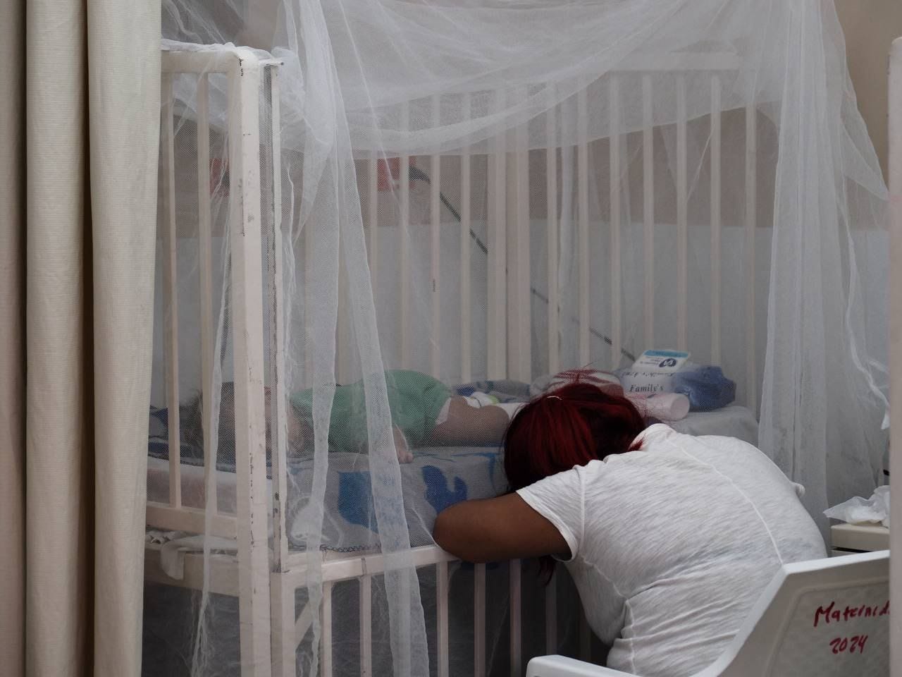 Mujer sentada frente a cama donde está paciente con dengue en hospital de honduras.